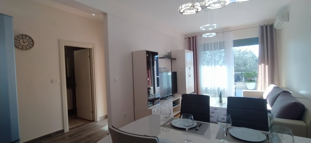 Modern living room with dining area in an apartment, Bečići, Montenegro: light furniture, TV area, and balcony access.