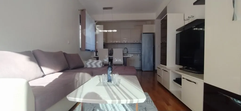 Living room with sofa, coffee table, and TV; kitchen visible in the background.