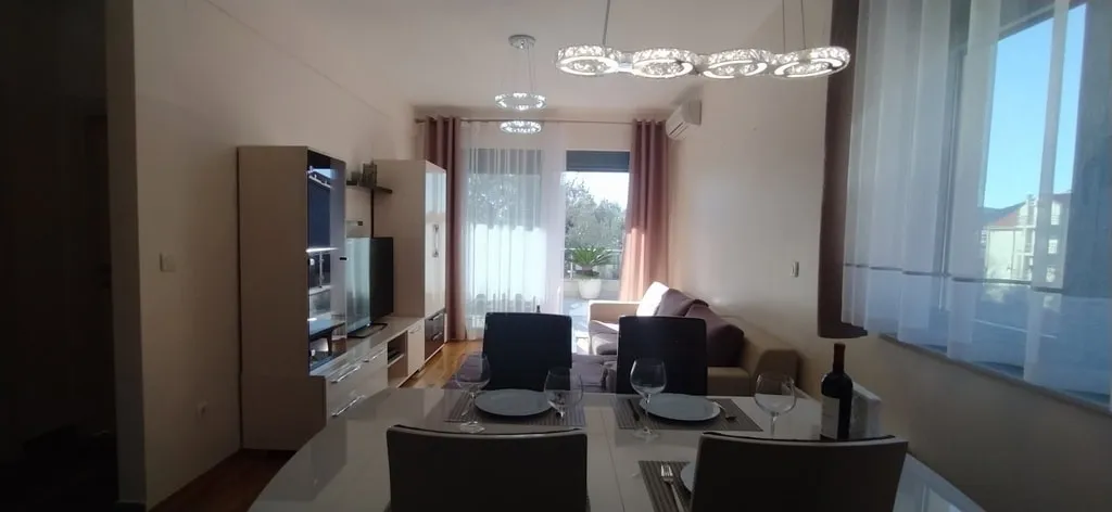 Living room with dining table, sofa, TV unit, chandelier, and large window with curtains.