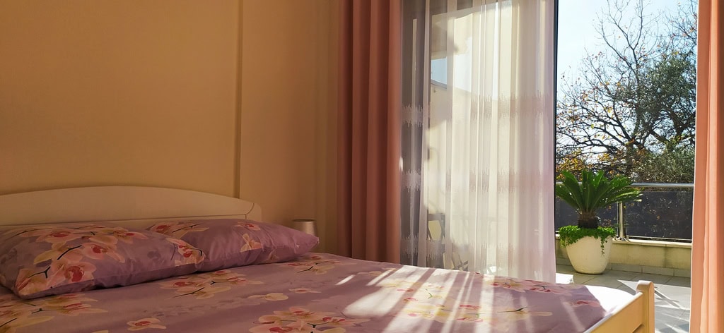 Bedroom in an apartment in Bečići, Montenegro: double bed, light interior, and access to a balcony with a garden view.