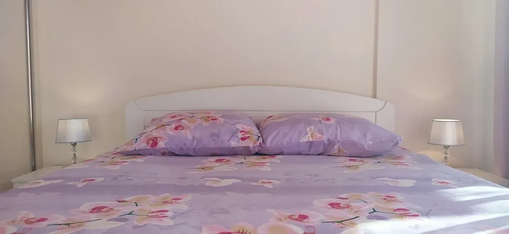 Bed with purple floral bedding and two lamps on bedside tables against a plain wall.