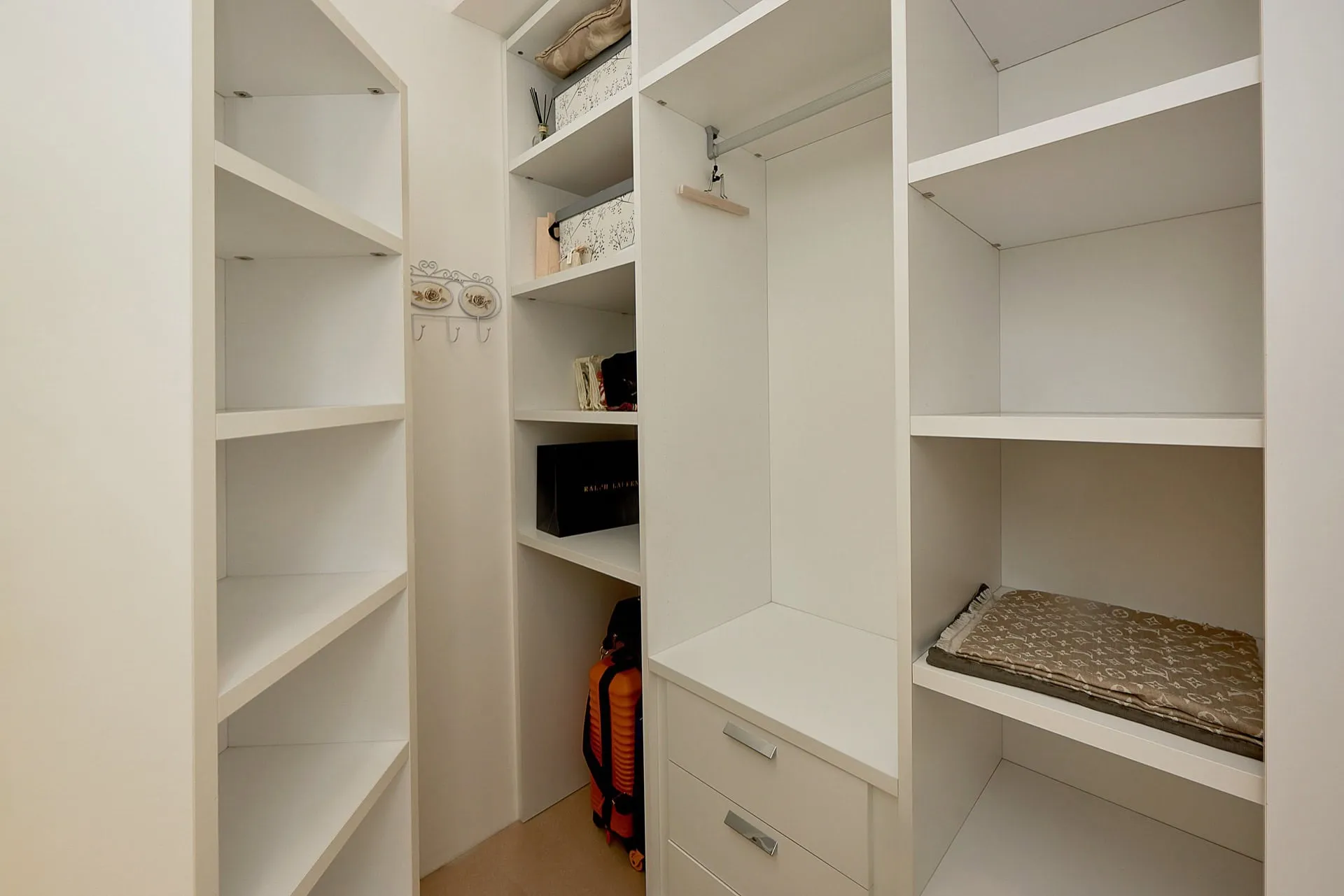 Built-in white walk-in closet with shelves, drawers, a hanging rod, and an orange suitcase on the floor.