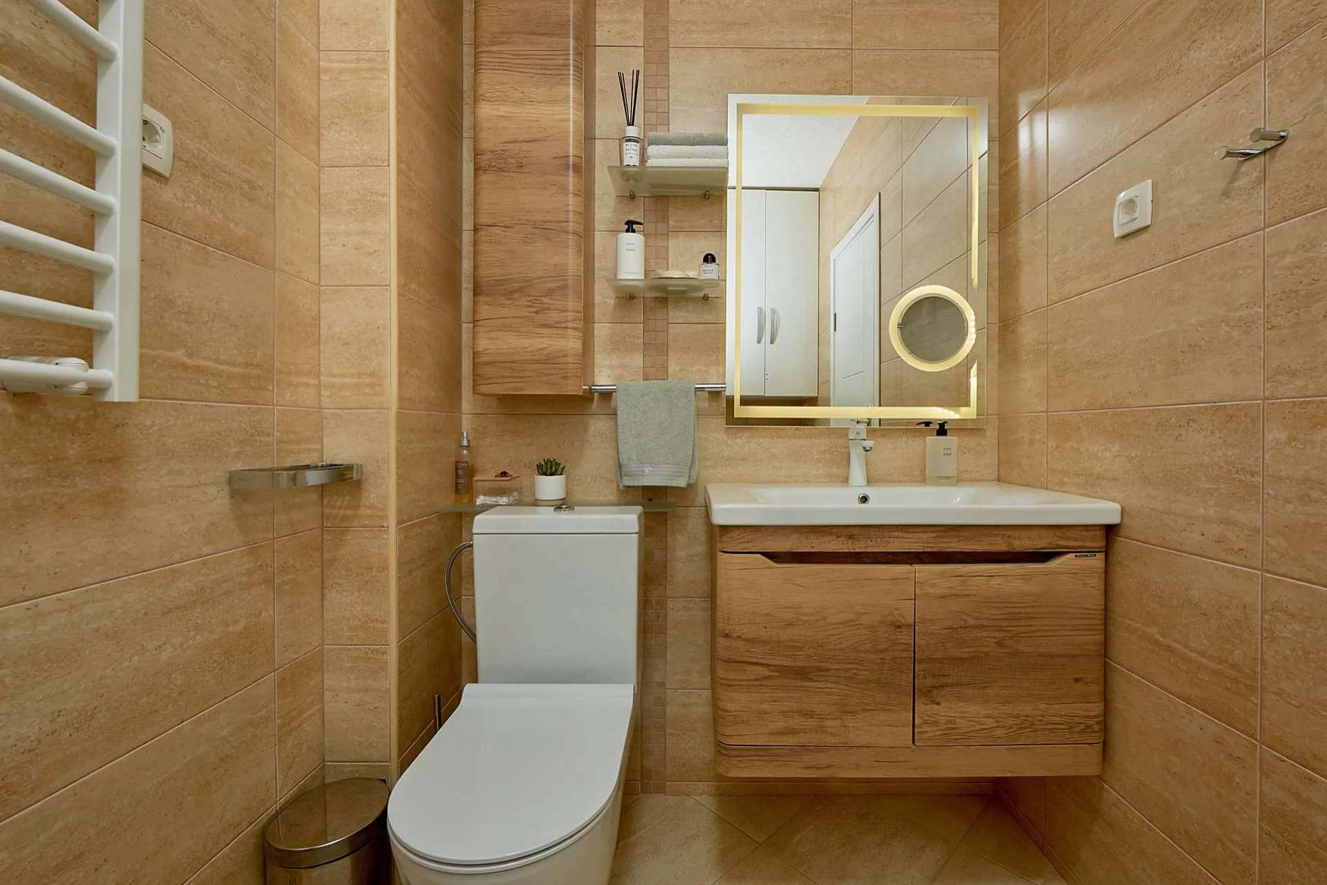 Bathroom featuring a toilet, wooden vanity sink, backlit mirror, and heated towel rail set against beige tiled walls.