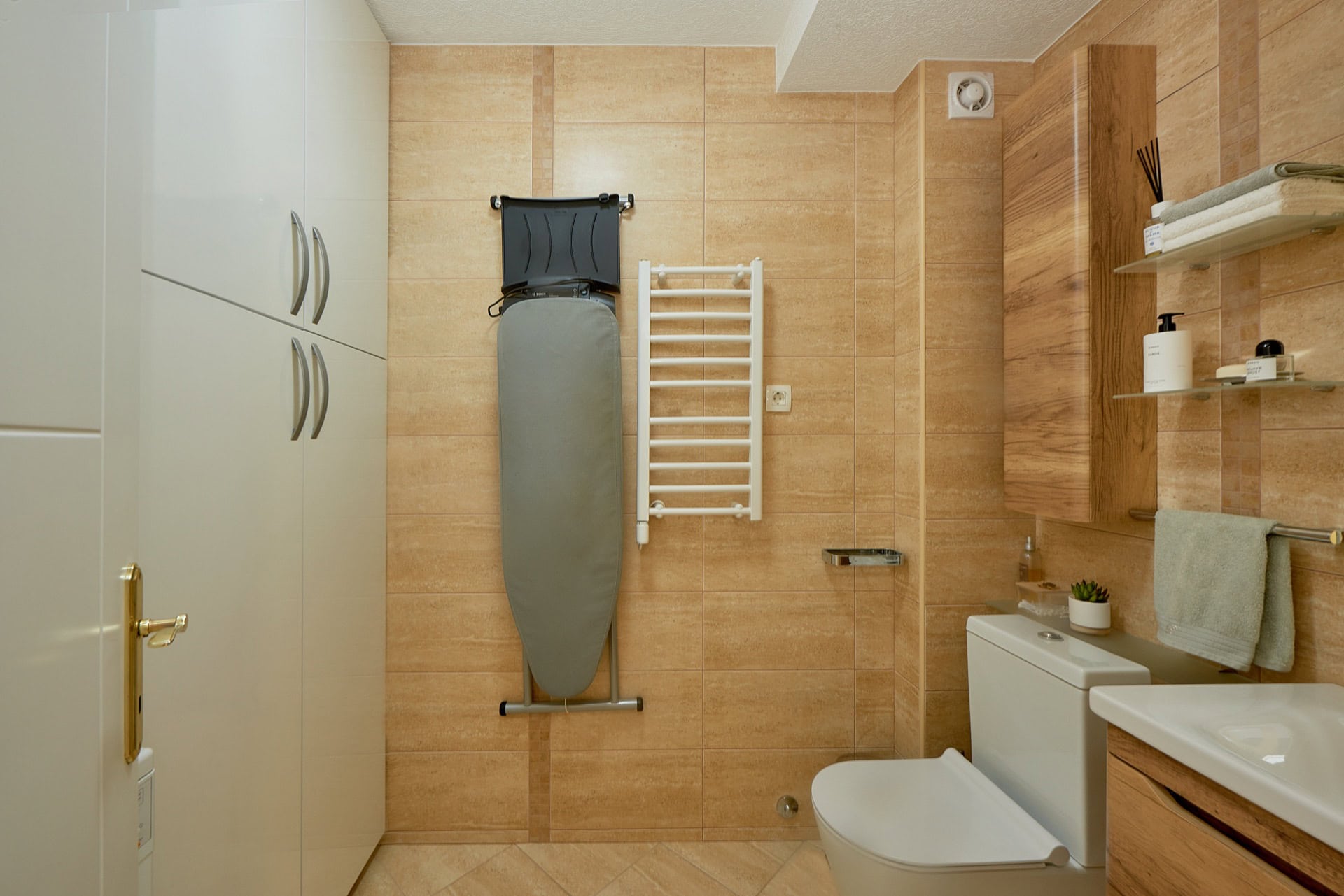 Bathroom in a penthouse in Bečići, Montenegro: beige tiles, built-in wardrobe, plumbing fixtures, and ironing board.