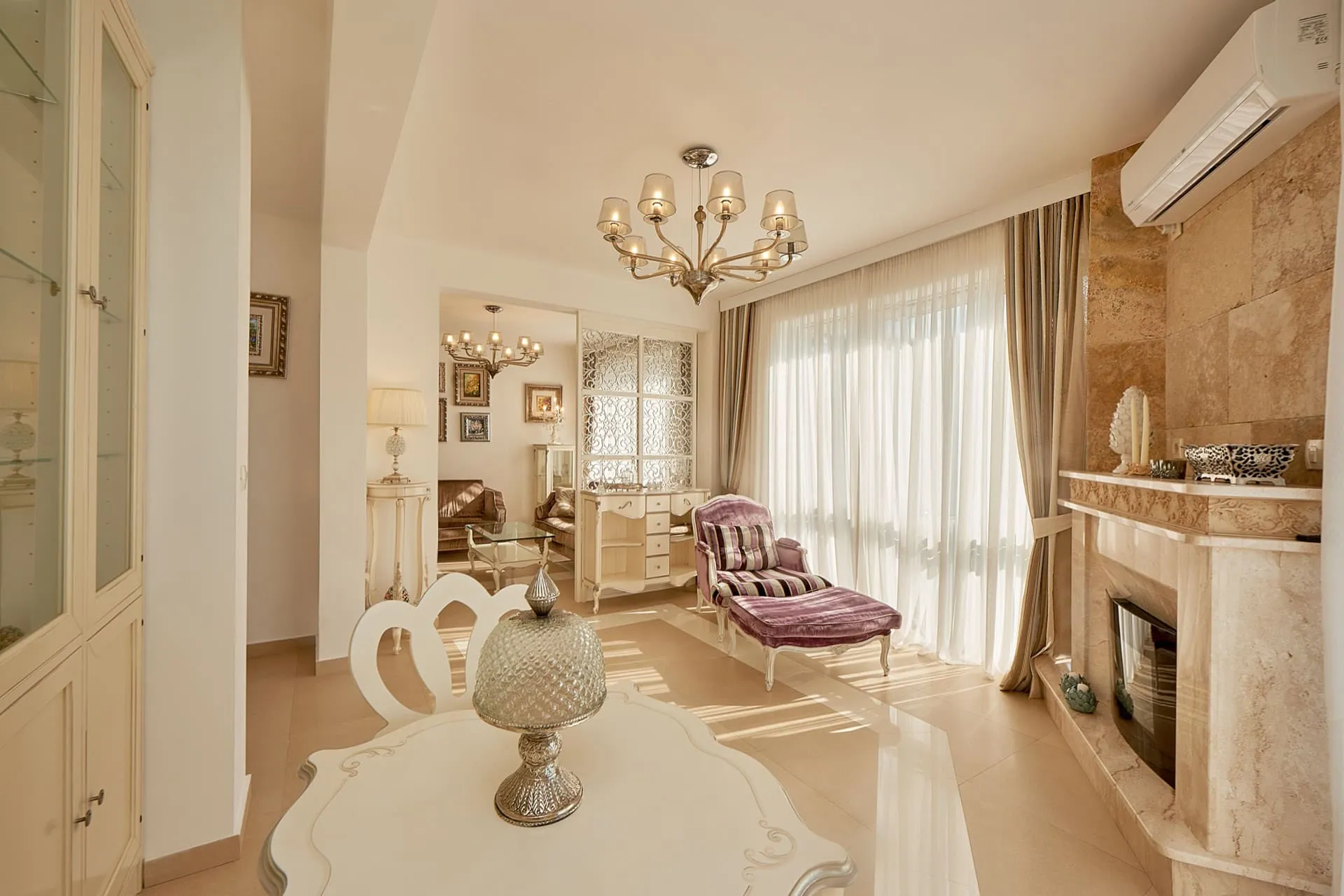 Bright living room featuring a stone fireplace, purple chaise lounge, chandelier, and white furniture.