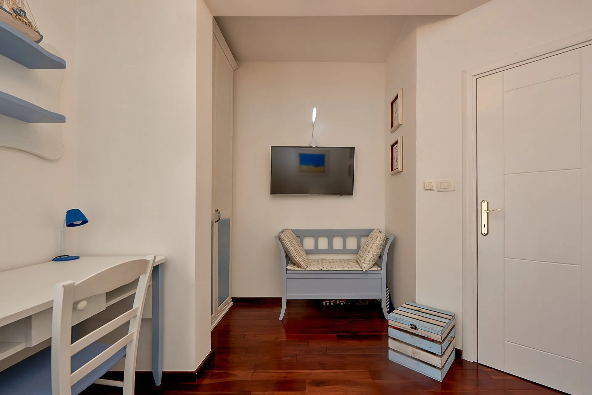Bright room with a white desk, blue chair, small sofa, and wall-mounted TV on dark wood floors.