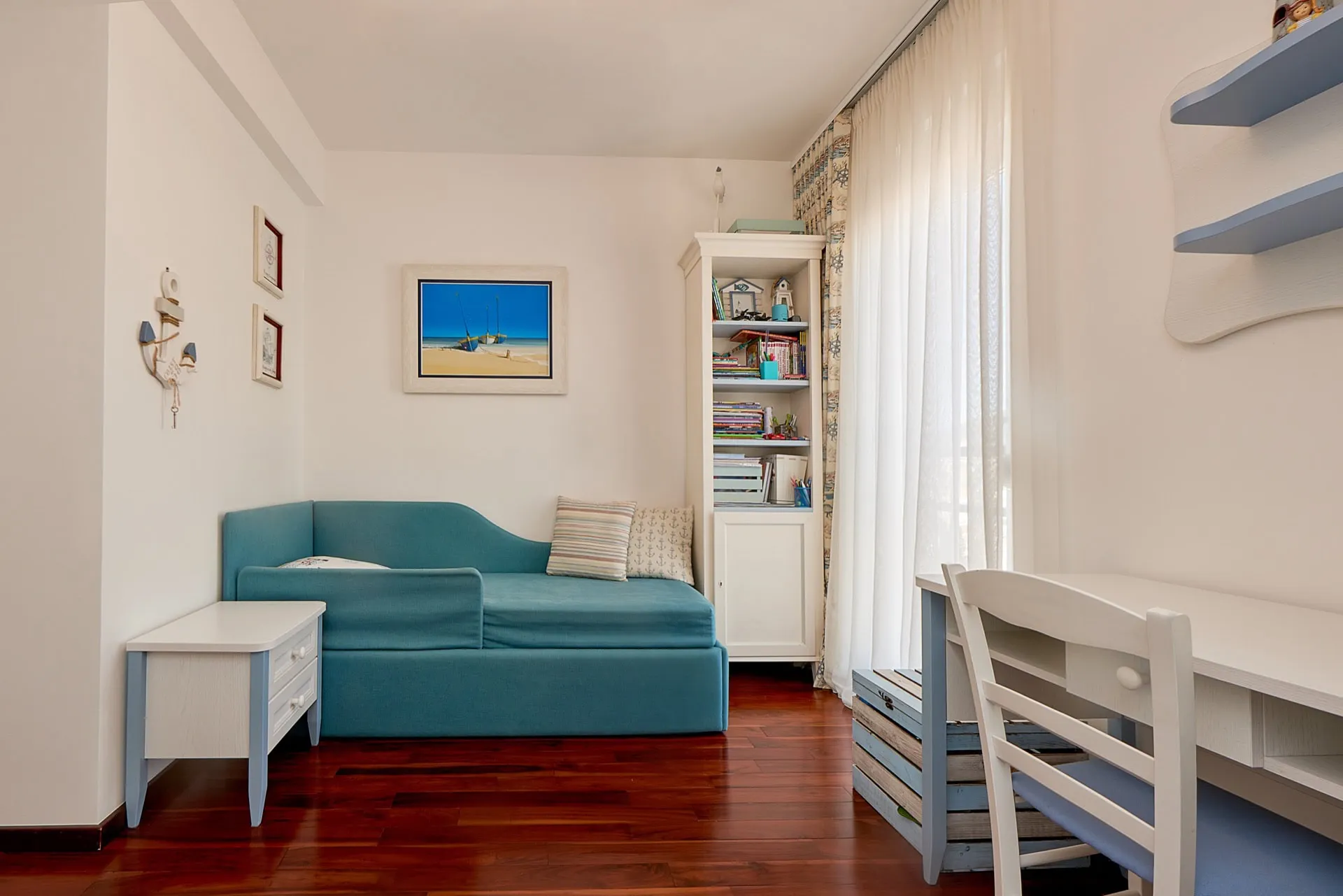 Blue daybed sofa, white desk, bookshelf, and nautical decor on dark hardwood flooring.