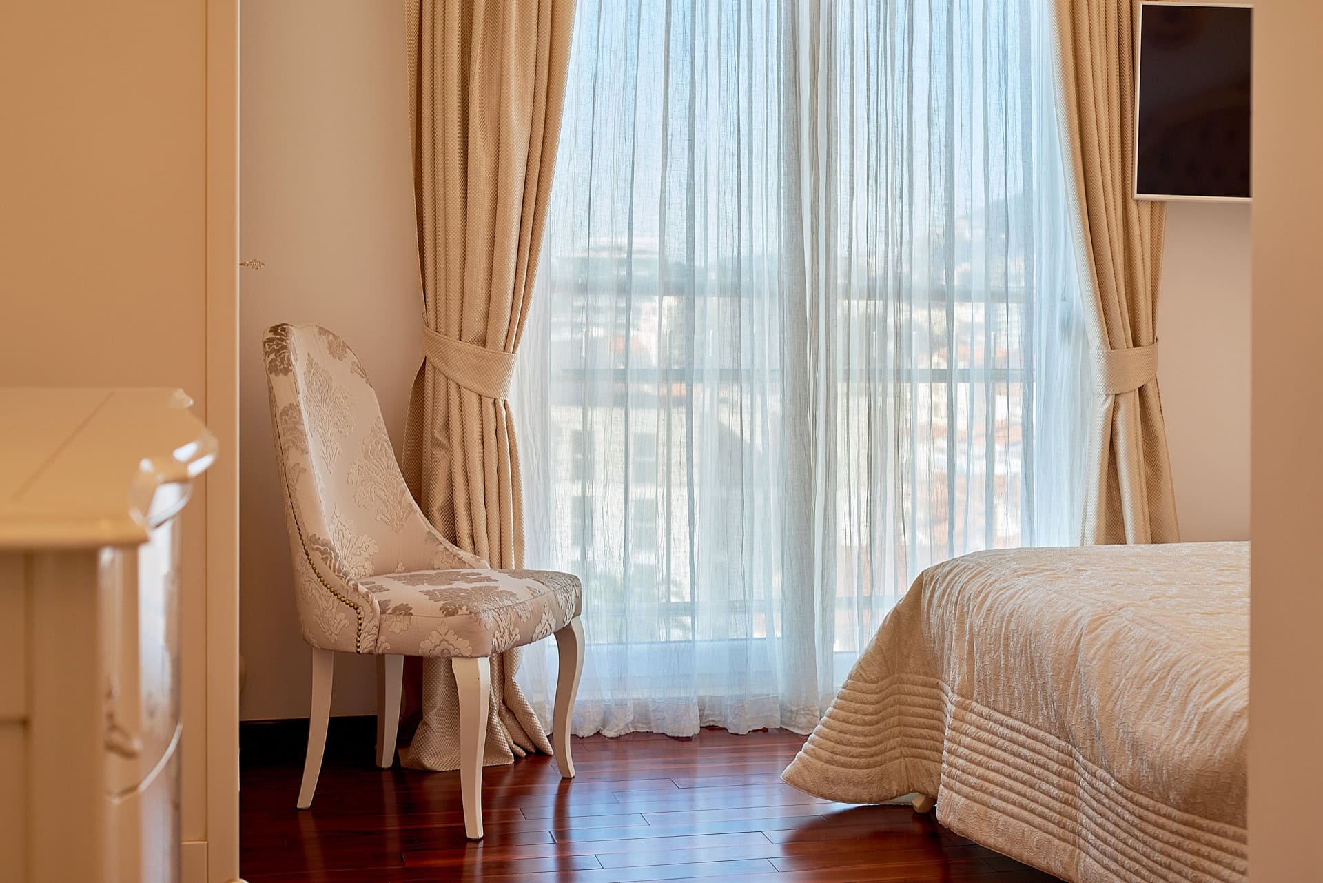 Bedroom in a penthouse in Bečići, Montenegro: classic interior, vintage chair, panoramic window with beige curtains.