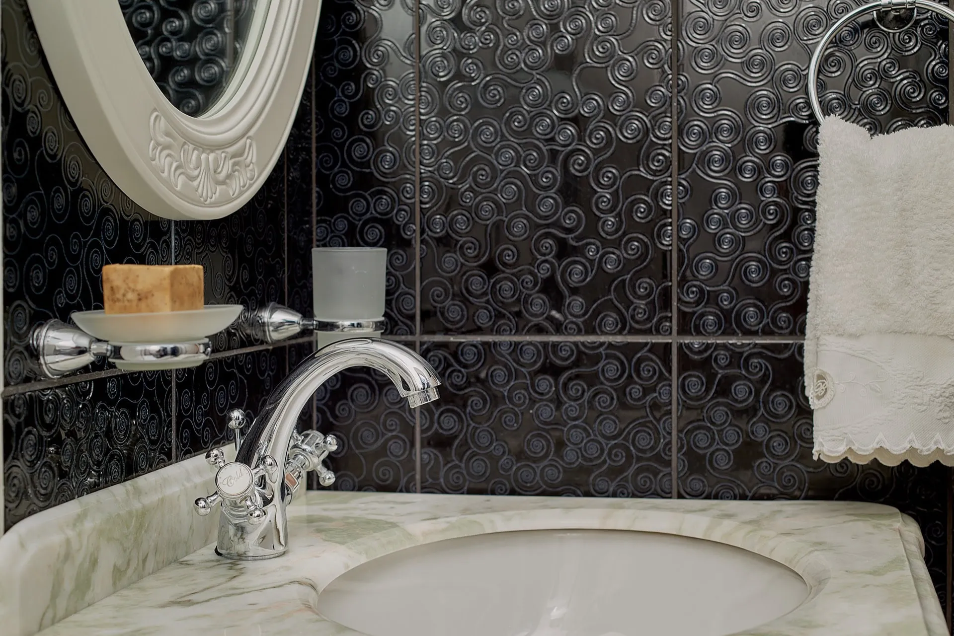 White marble sink with chrome faucet, black tiles with swirl pattern, oval mirror, and white towel on a ring.