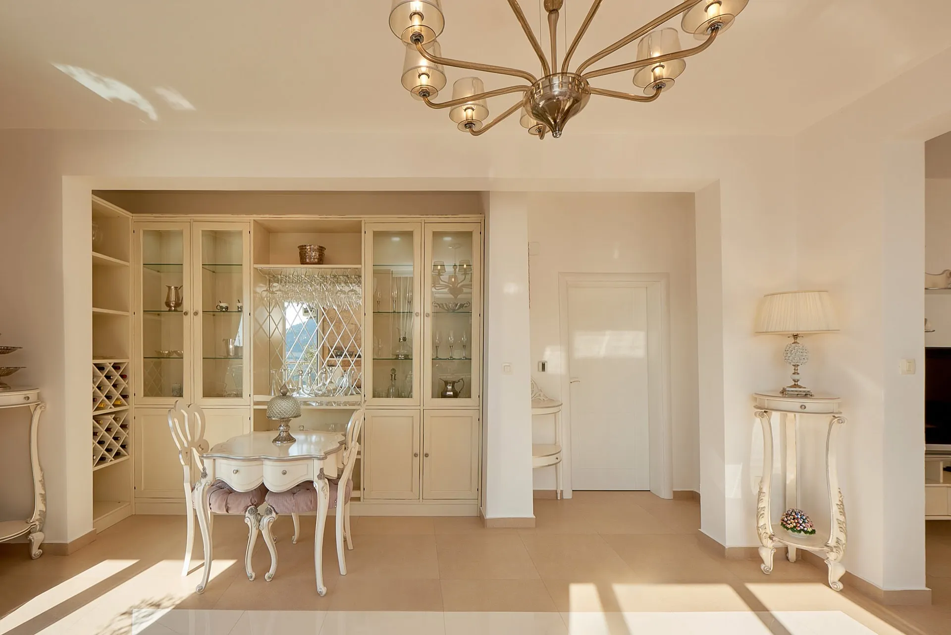 Bright room featuring a dining area with a white glass-front cabinet, wine rack, ornate table, and chandelier.