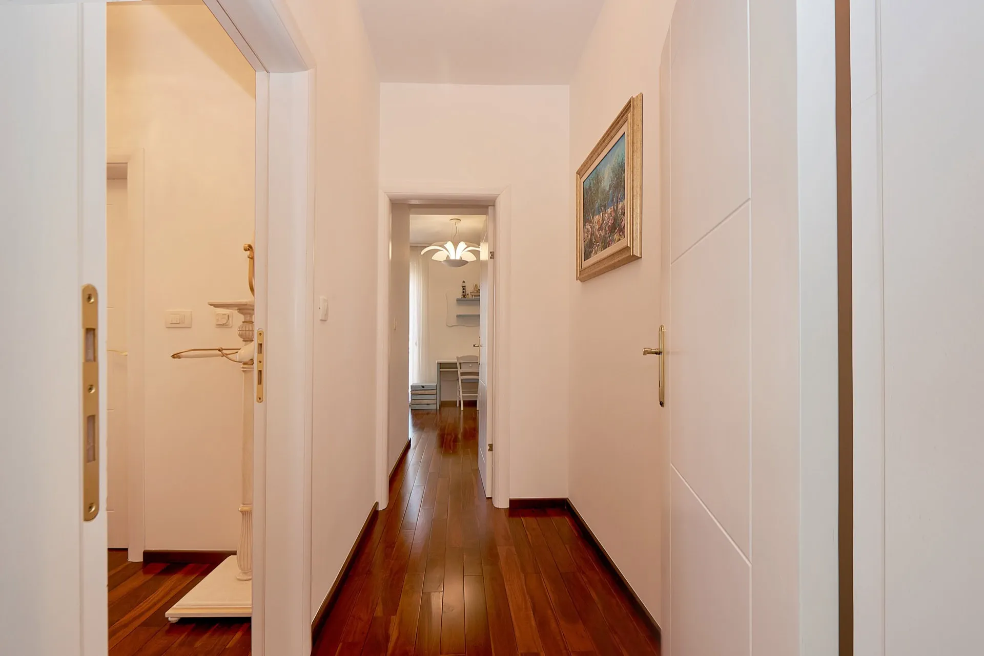 Long hallway with dark wood floors, white walls, a framed painting, and open doorways leading to other rooms.