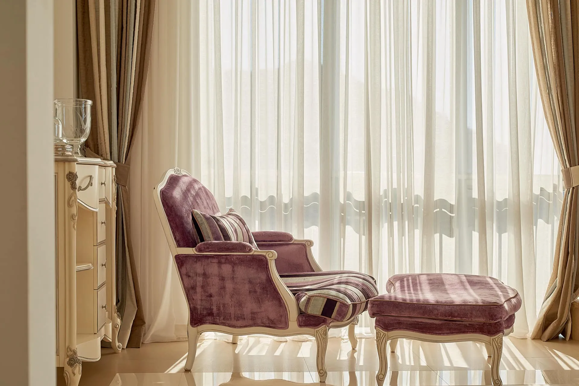 Classic purple armchair and ottoman by a window with sheer curtains, next to an ornate cream cabinet.