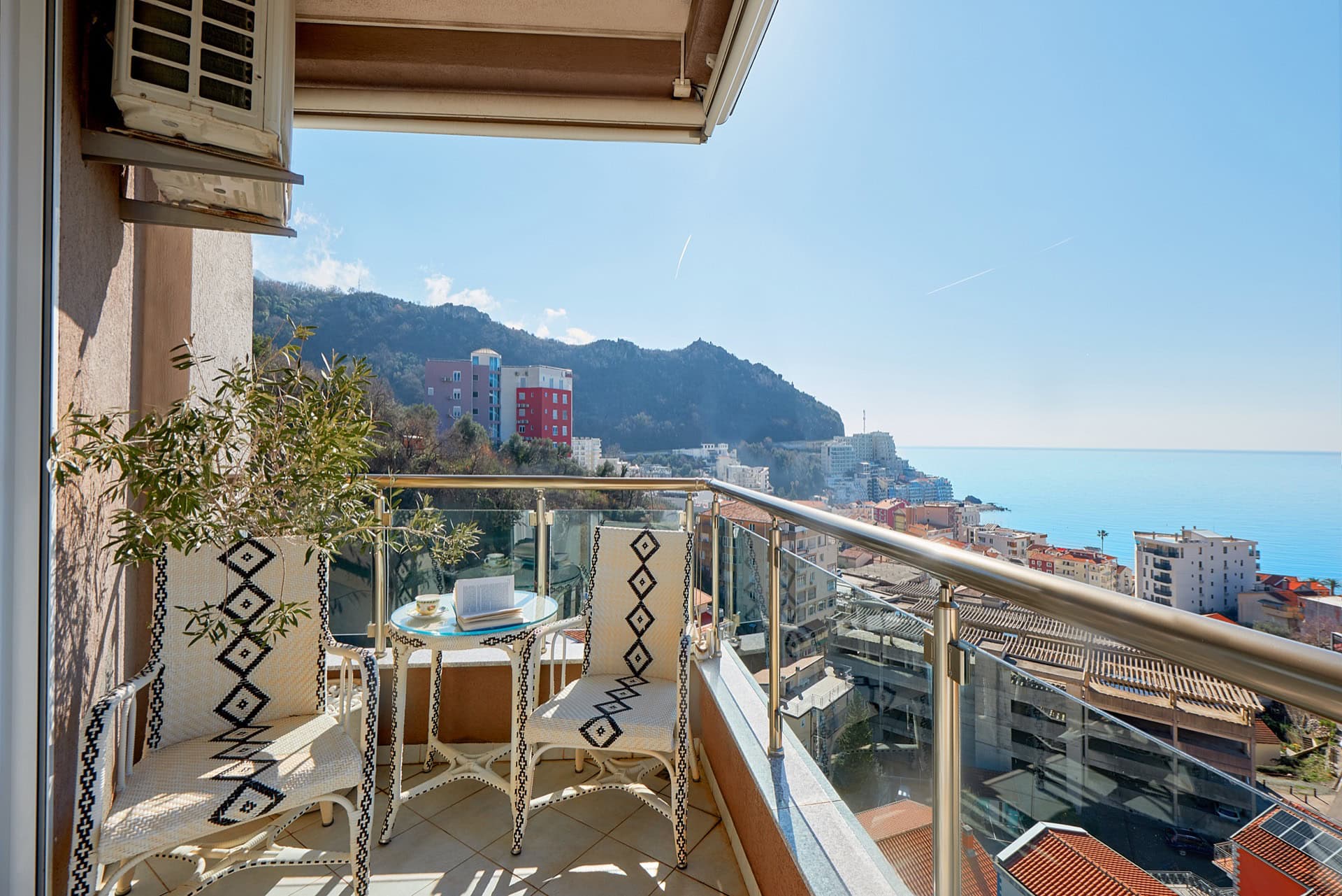 Penthouse balcony in Bečići, Montenegro: wicker furniture, panoramic view of the sea, mountains, and urban development.