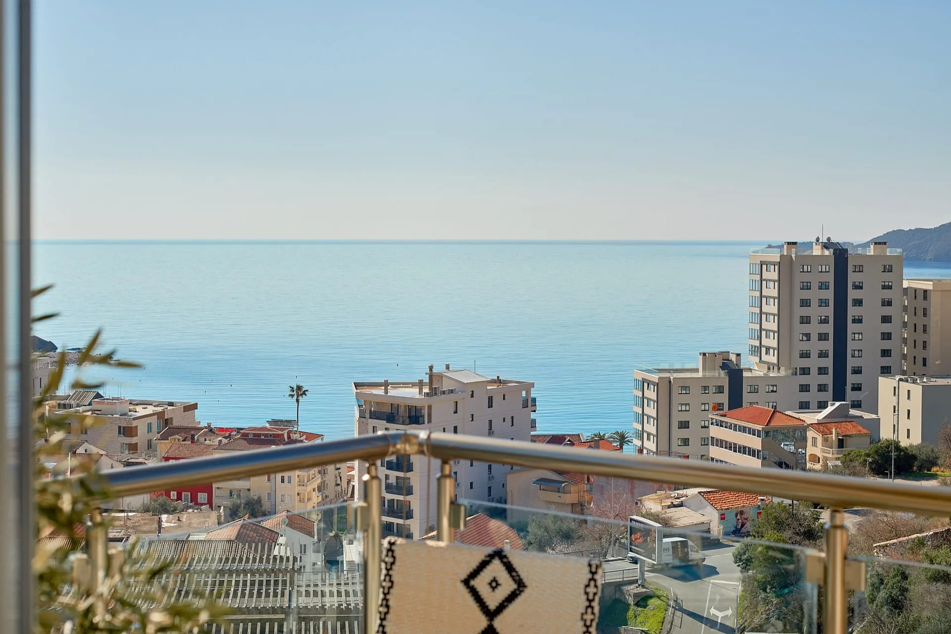 View from a balcony overlooking the sea and city buildings. A metal railing with glass panels and part of a chair are visible in the foreground.