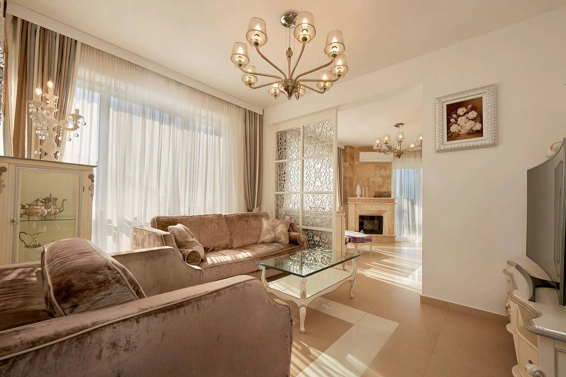 Bright living room with velvet sofas, glass coffee table, chandelier, and a fireplace visible in the adjacent area.