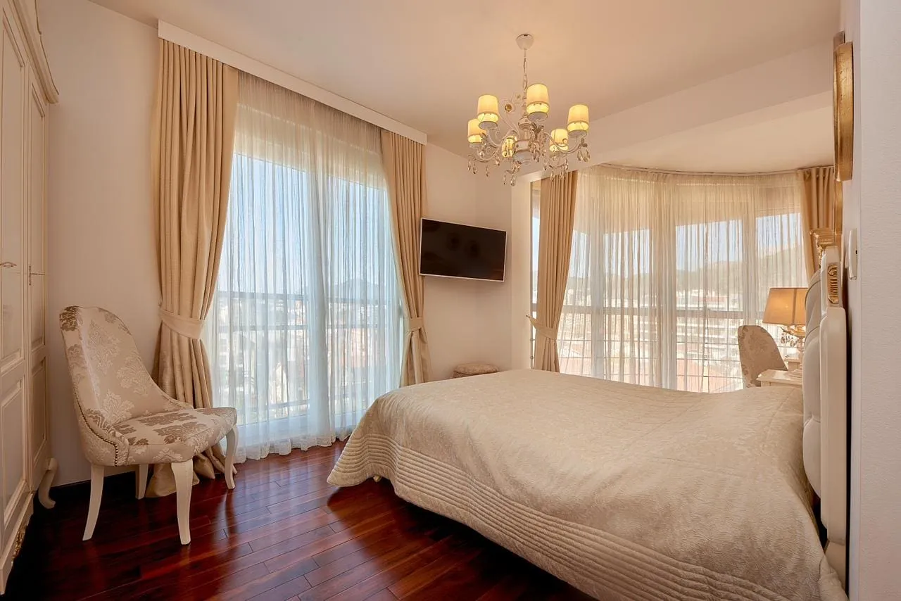 Bright bedroom featuring a large bed, crystal chandelier, dark wood floors, and an upholstered chair.