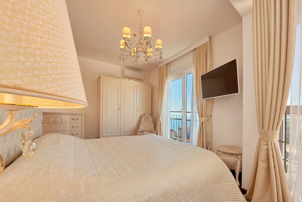Bright bedroom featuring a large bed, white wardrobe, chandelier, and wall-mounted TV near the window.