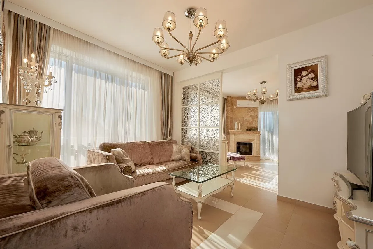 Living room with velvet sofas, glass coffee table, chandelier, and a fireplace visible in the background.