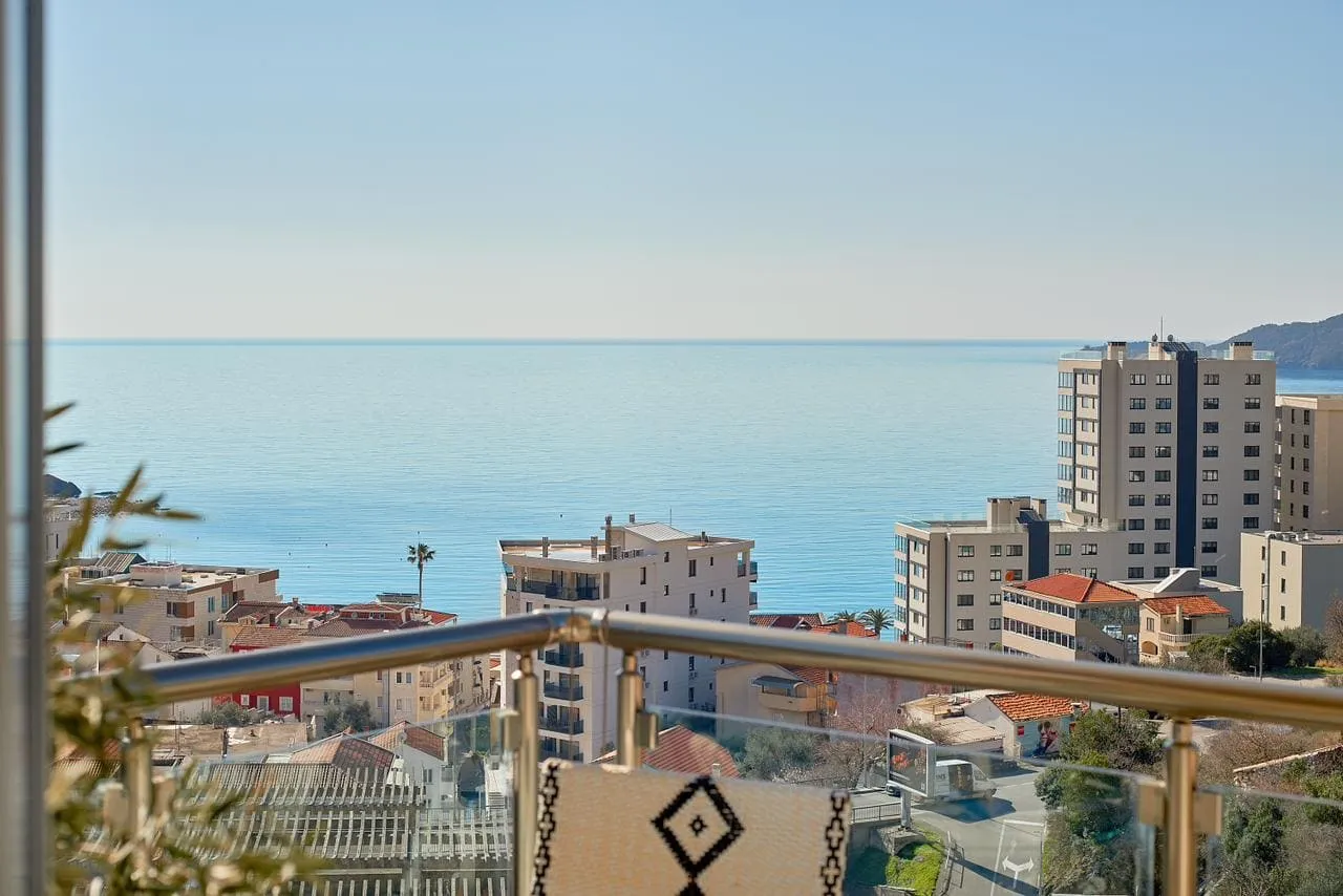 View from a balcony overlooking the sea and city buildings through a glass railing with a metal handrail.