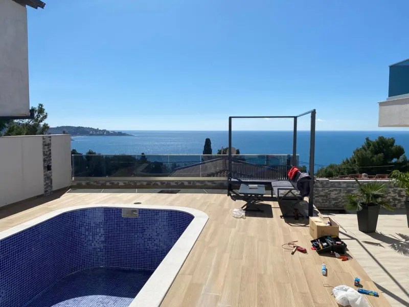 Blue tiled pool on a wooden deck with sea view, tools and furniture present nearby.