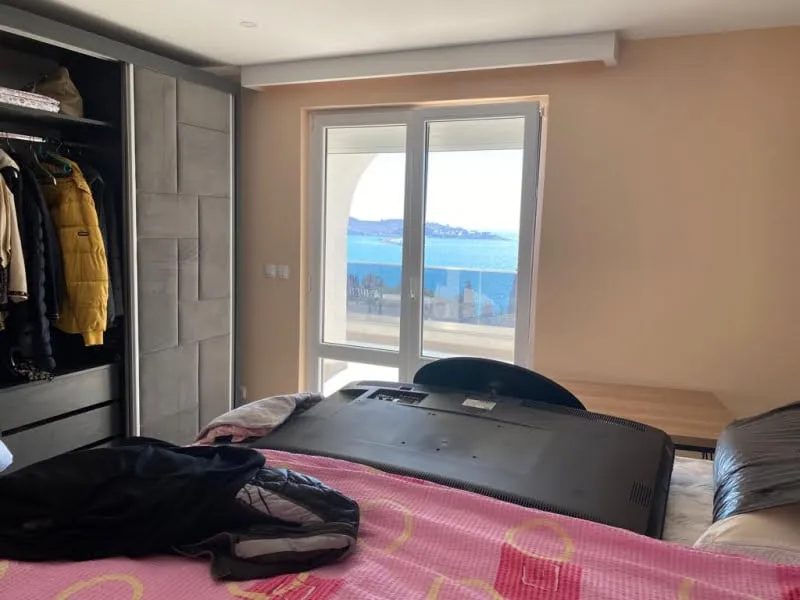 Bedroom with a pink bedspread, open wardrobe with clothes, and a TV on the bed facing a window.