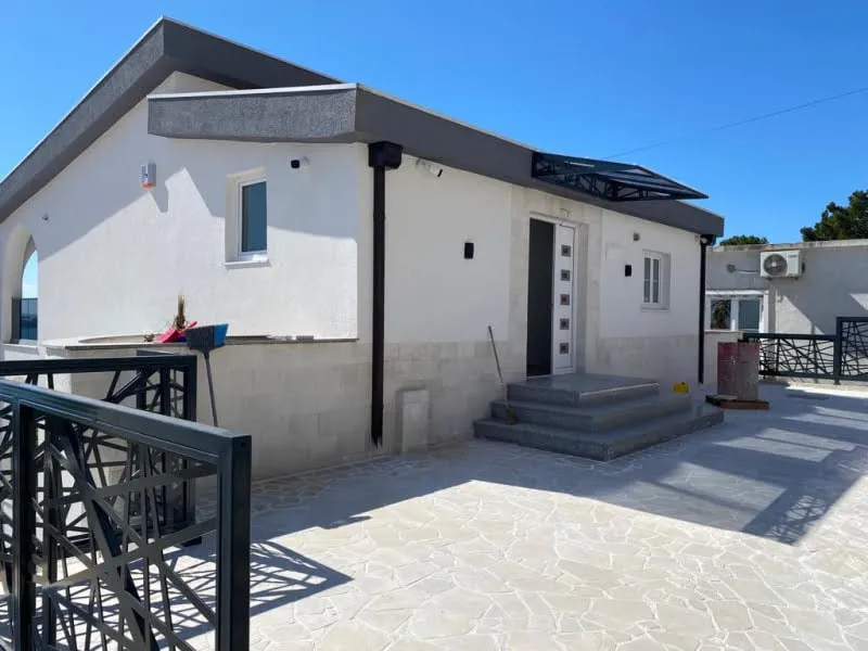 Exterior view of a white single-story house with a stone patio, entrance steps, and black metal railing under a clear blue sky.