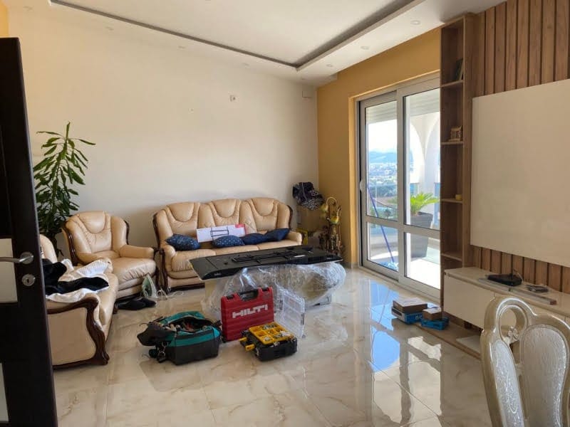 Living room with beige leather furniture, tools on the floor, and a sliding glass door to a balcony.