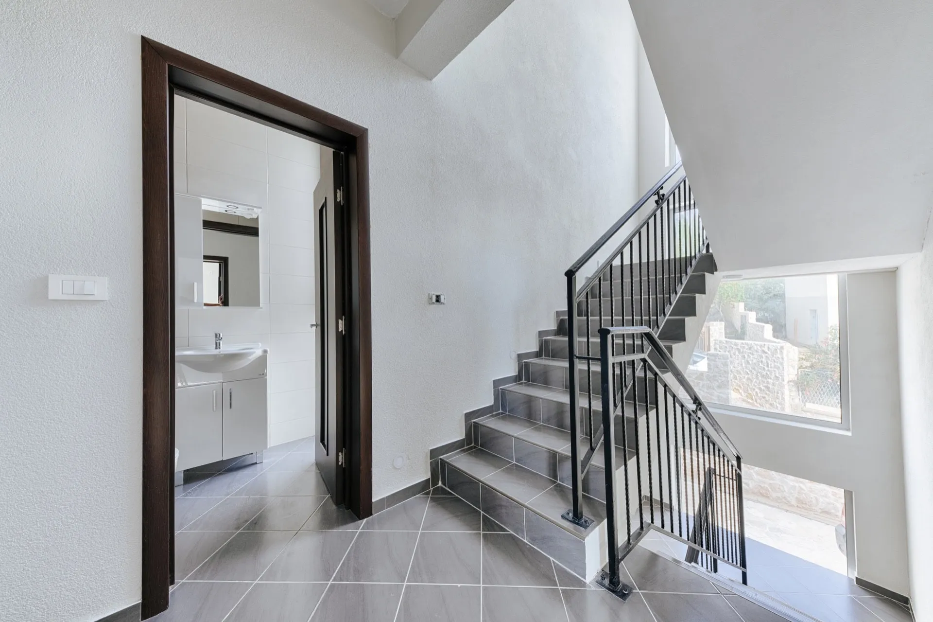 Entryway with gray tile flooring, a staircase with black metal railing, and an open door revealing a bathroom vanity inside.