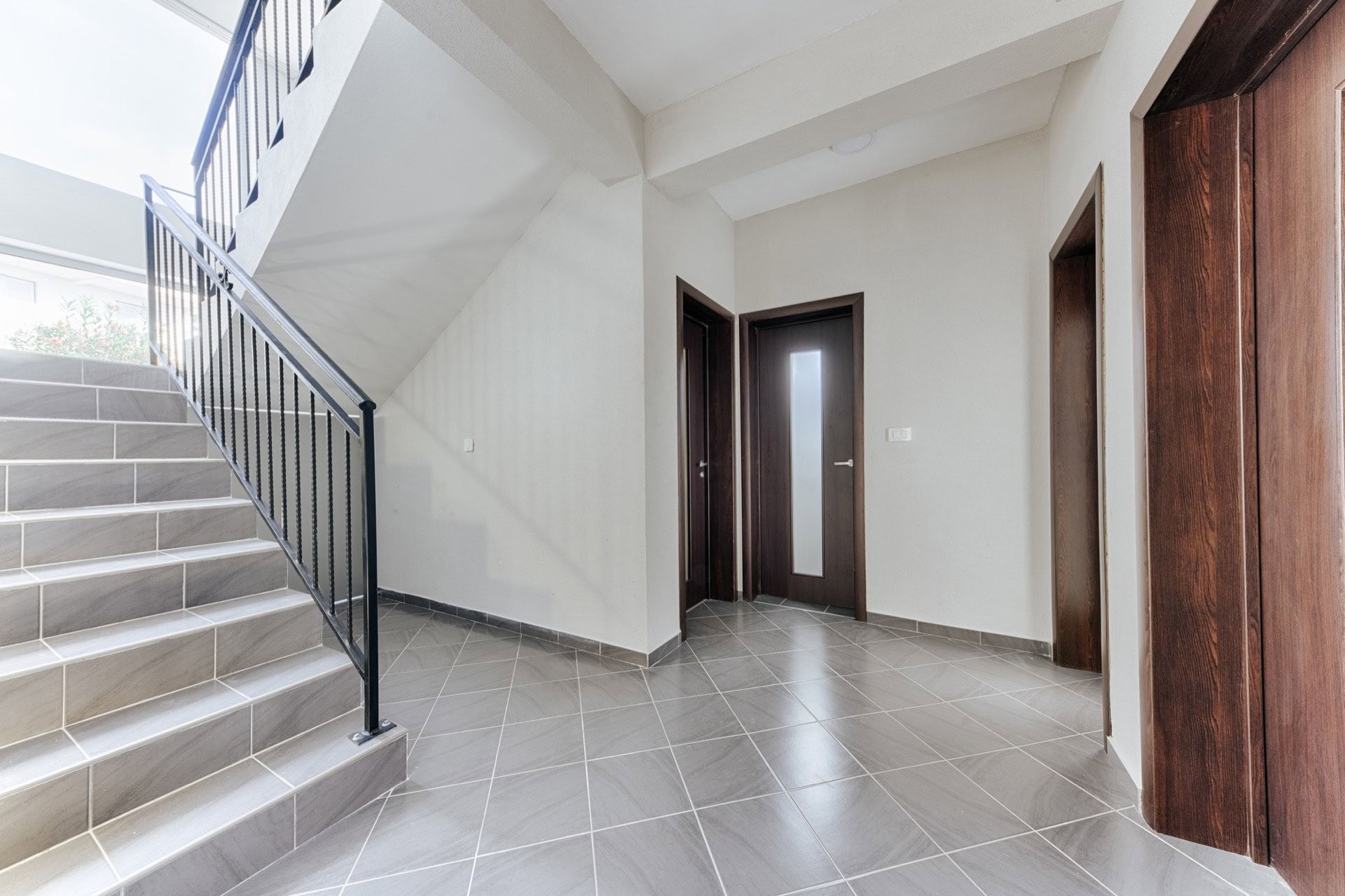 Staircase with wrought-iron railings and a bright villa hall in Utjeha, Montenegro, with gray tile finishes and dark doors.
