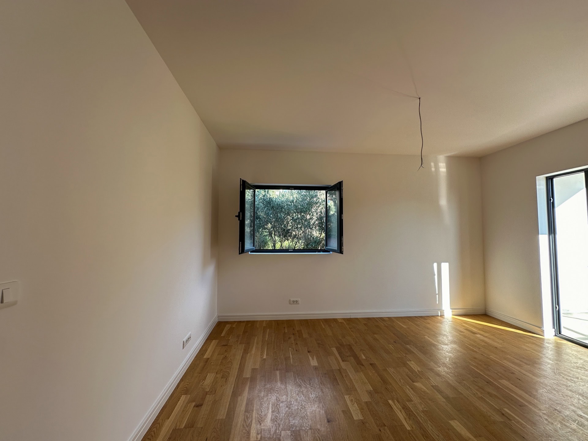 Bright living room with parquet flooring in a new apartment in Bečići, Montenegro, with a view of greenery from the open window.