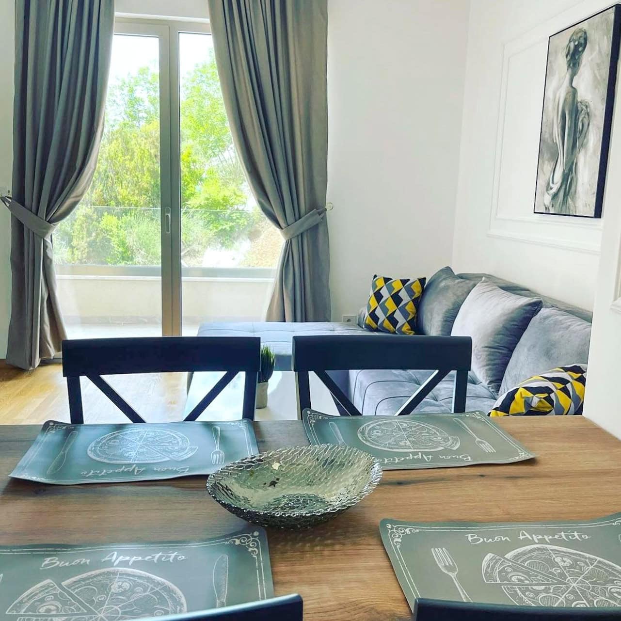 Living room with dining area in an apartment in Bečići, Montenegro: gray sofa, wooden table, and a view of greenery from the window.