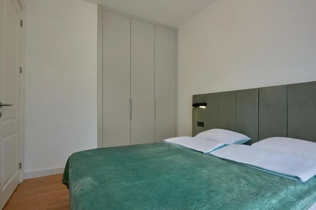 Bedroom with a double bed covered in green fabric and a grey upholstered headboard against white walls.