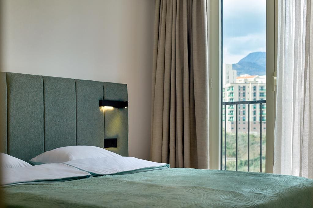 Bedroom in an apartment in Bečići, Montenegro: a double bed with a soft headboard and mountain views from a panoramic window.