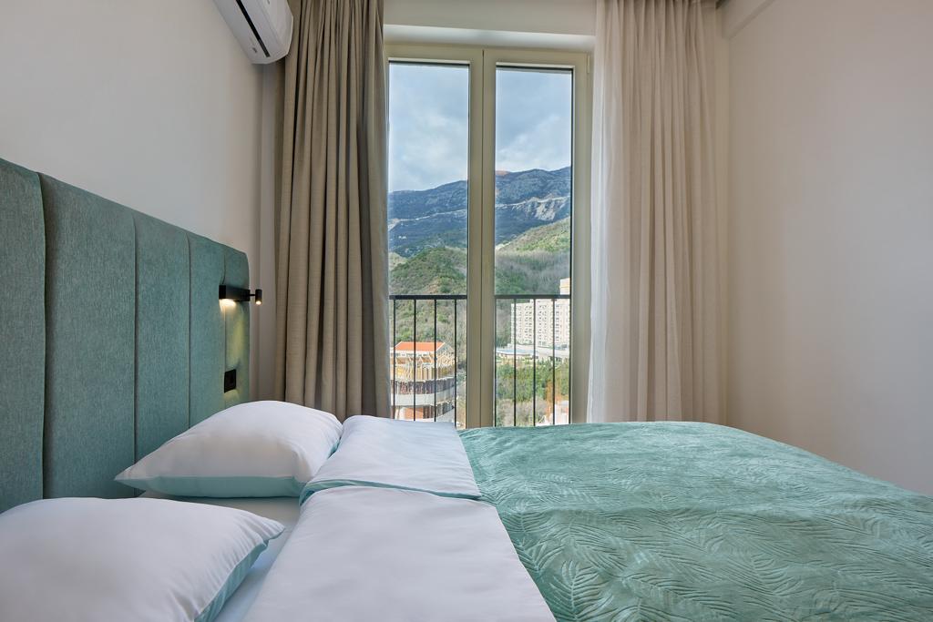 Modern bedroom in an apartment in Bečići, Montenegro: double bed, mountain view, and a balcony with a panoramic window.