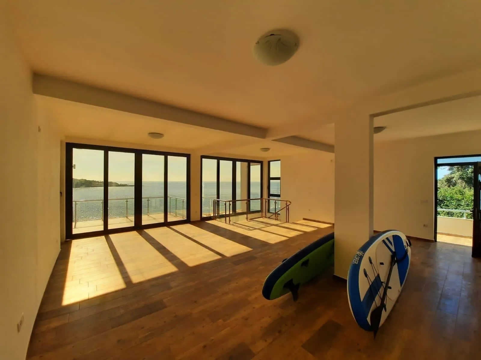 Empty room with wooden floors and large windows overlooking the sea; two paddleboards lean against a wall.