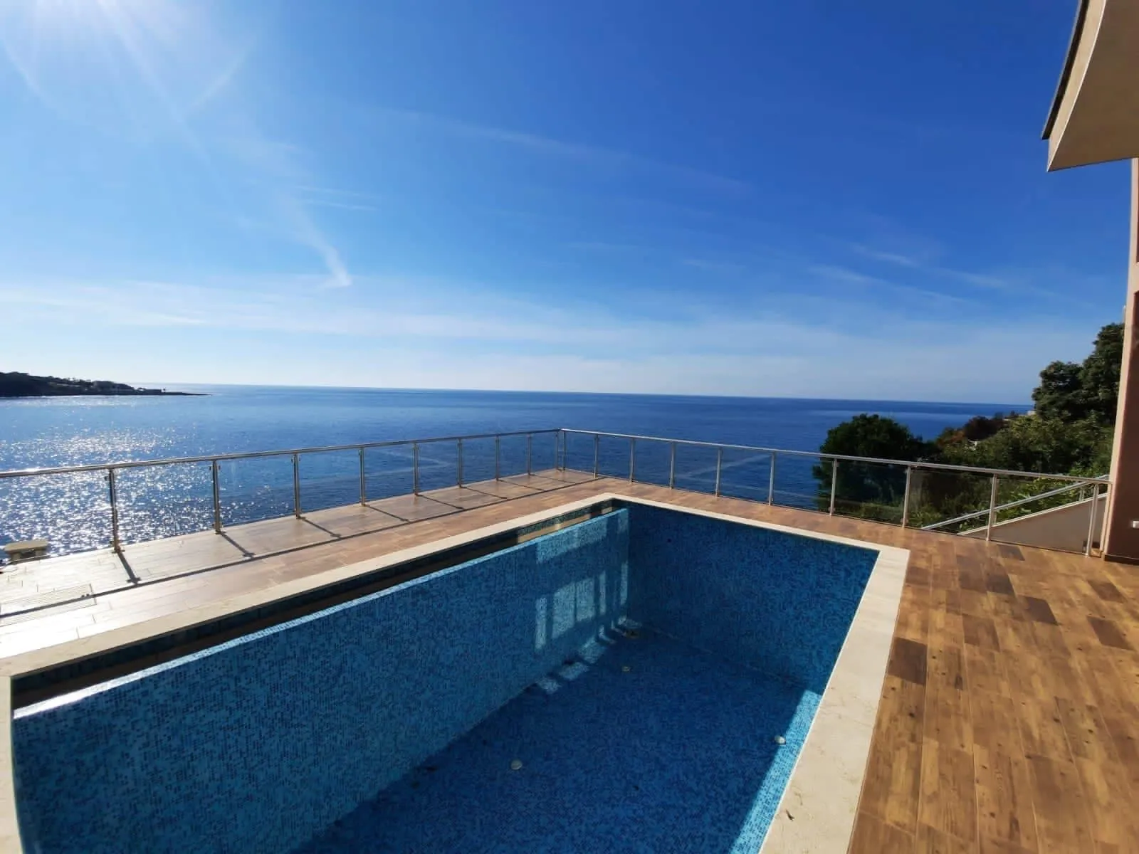 Empty blue-tiled swimming pool on a wooden deck terrace overlooking the sea under a clear sky.