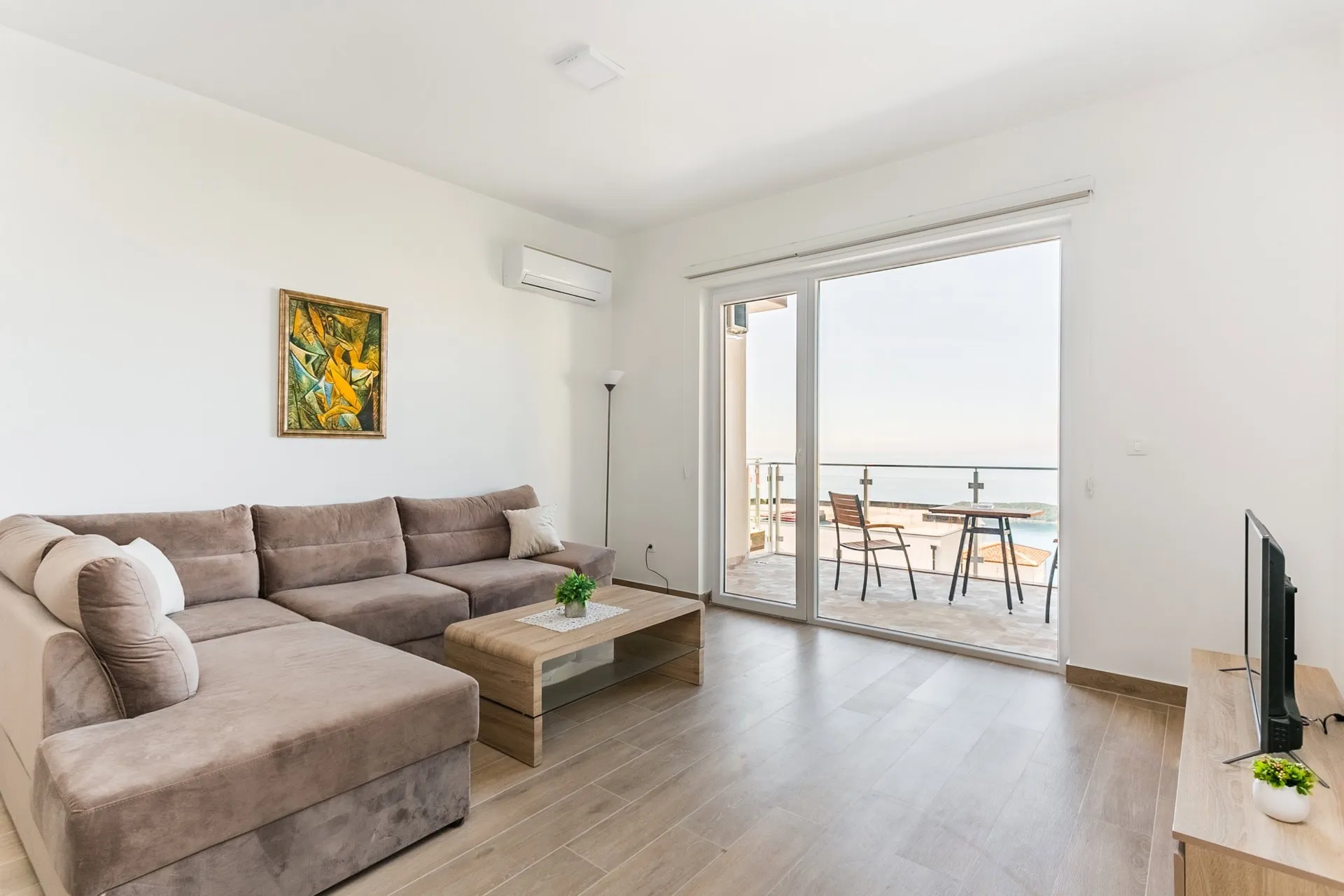 Bright living room with a grey sectional sofa, wooden coffee table, and TV. Sliding glass door leads to a balcony with a sea view.