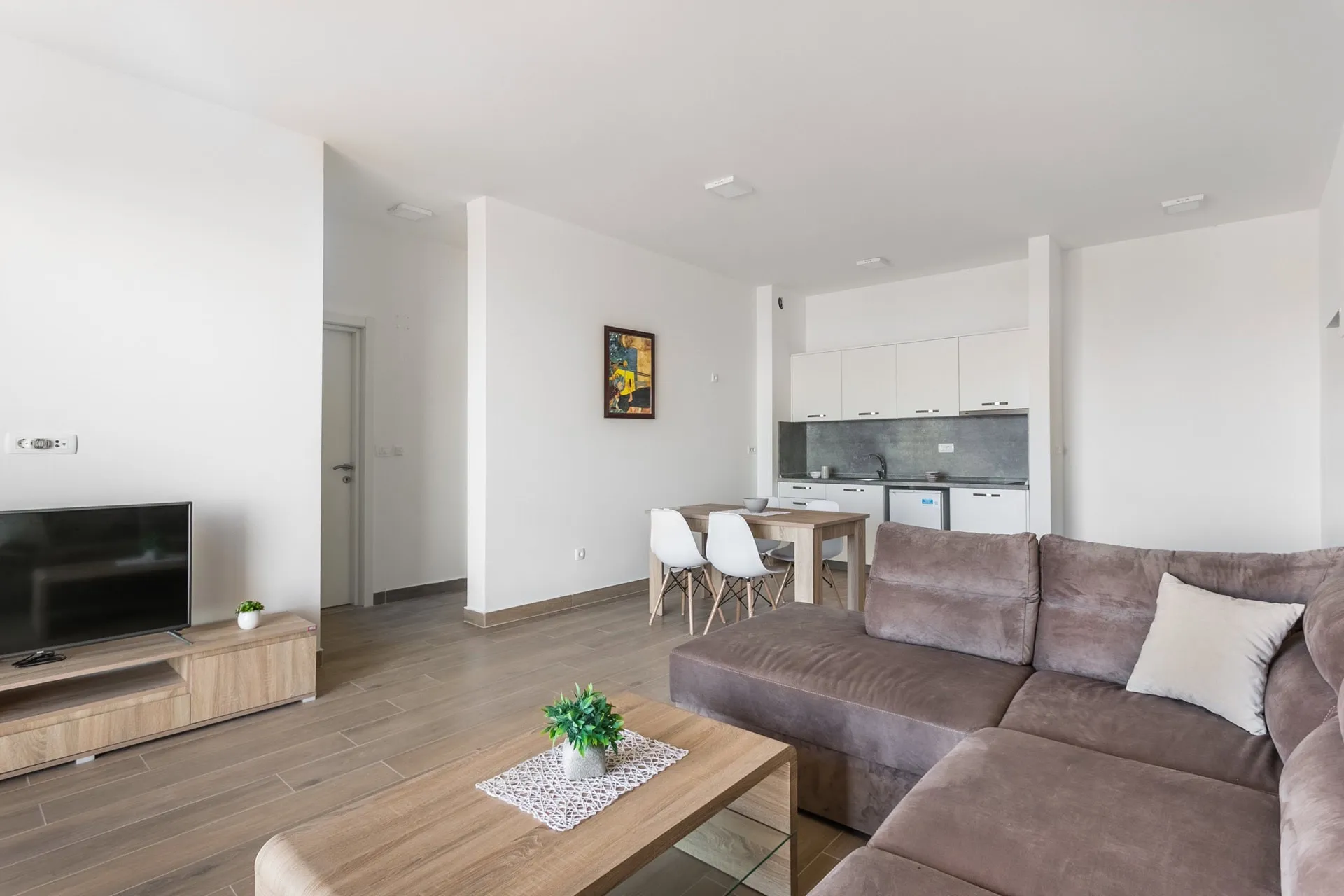 Bright living room with a grey sofa, wooden dining table, and an open kitchen area visible in the background.