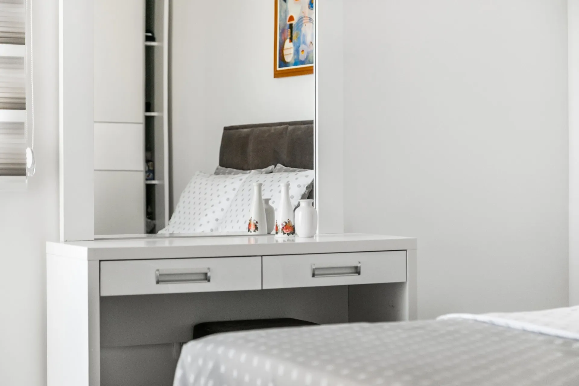 White vanity table with mirror and decorative vases; a bed with a brown headboard is visible in the reflection.