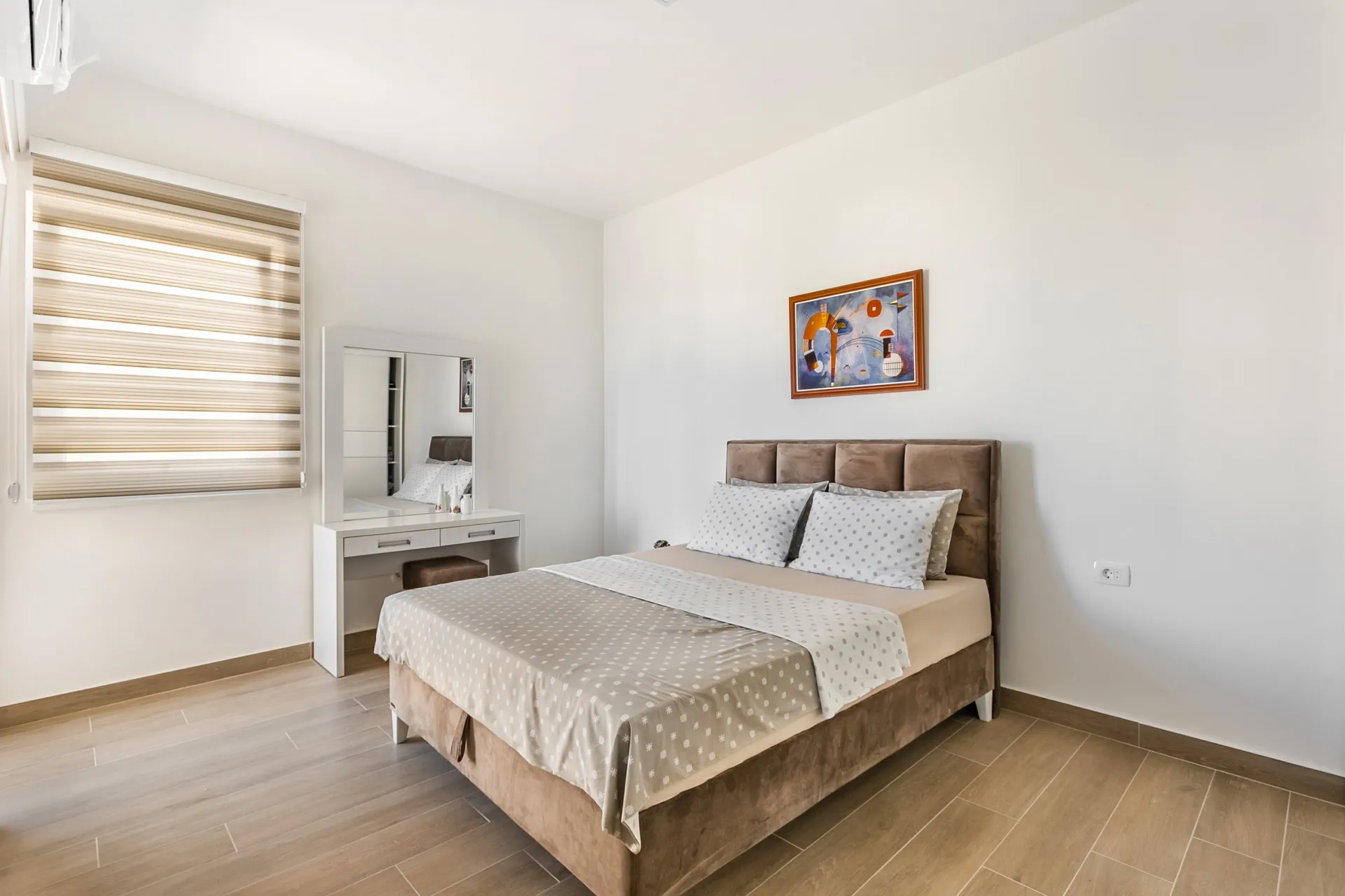 Bright bedroom featuring a double bed with brown headboard, white vanity table with mirror, and abstract wall art.