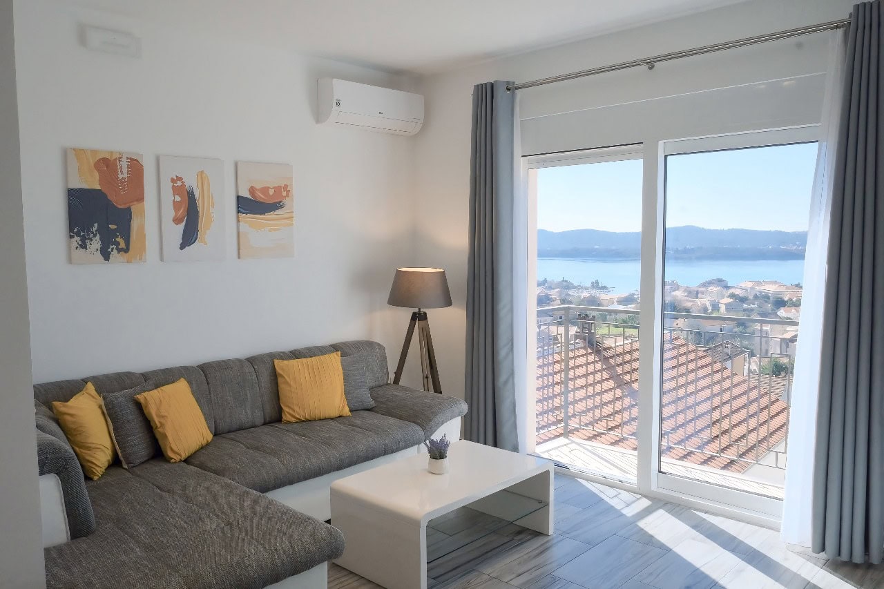 Bright living room in an apartment in Tivat, Montenegro: gray sofa, panoramic sea view, and a balcony with a terracotta roof.