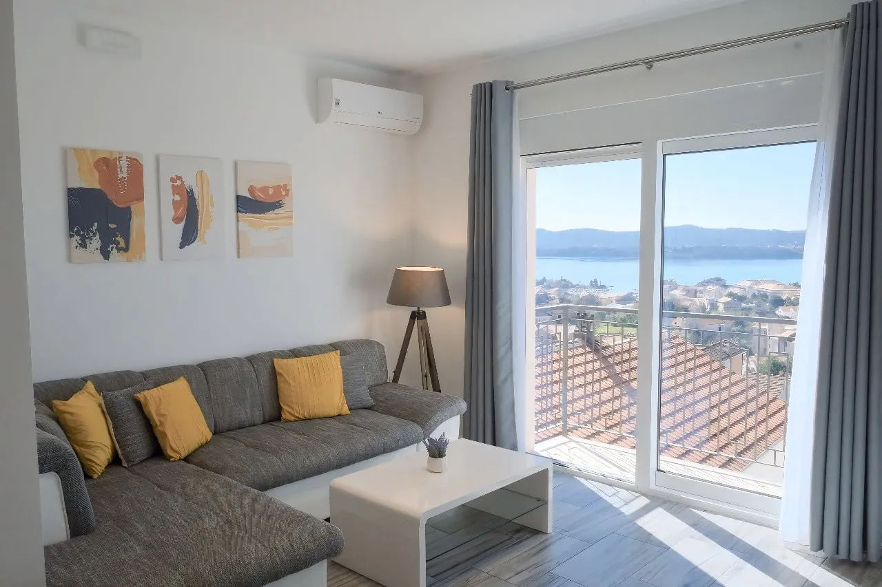 Living room with a grey sofa, white coffee table, and abstract wall art. Large glass door leads to a balcony overlooking the sea.
