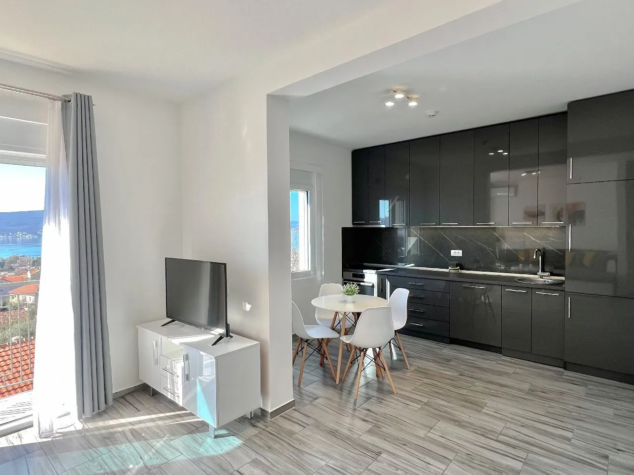 Open plan space with TV on white console and kitchen area featuring dark cabinets and a small dining table.