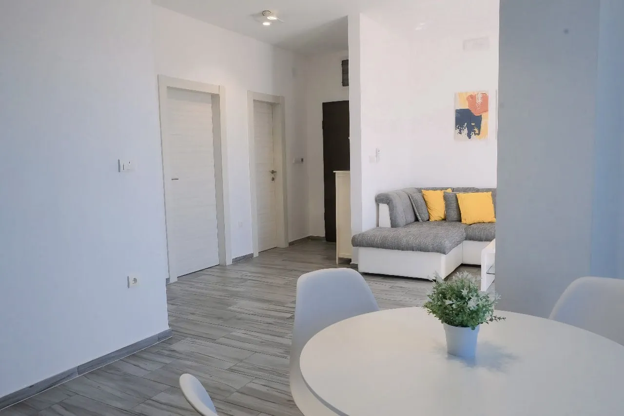Living area featuring a grey sectional sofa with yellow pillows and a white round dining table in the foreground.