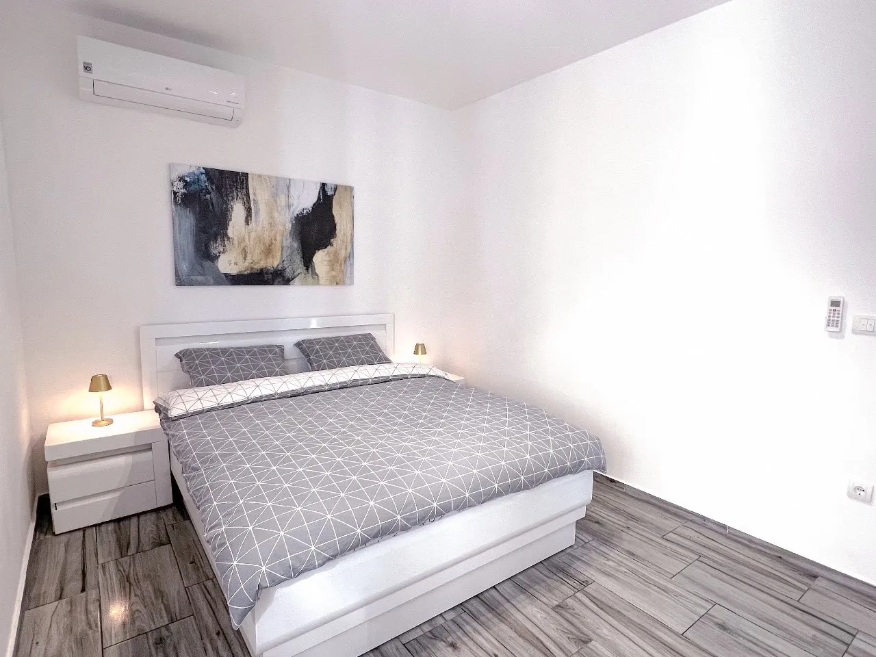 Modern bedroom featuring a white bed with grey geometric bedding, abstract wall art, and a wall-mounted air conditioner.