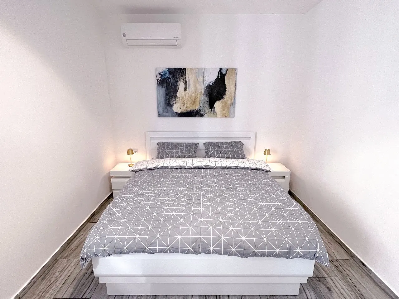 Modern bedroom with a white bed, grey geometric bedding, abstract wall art, and a wall-mounted AC unit.
