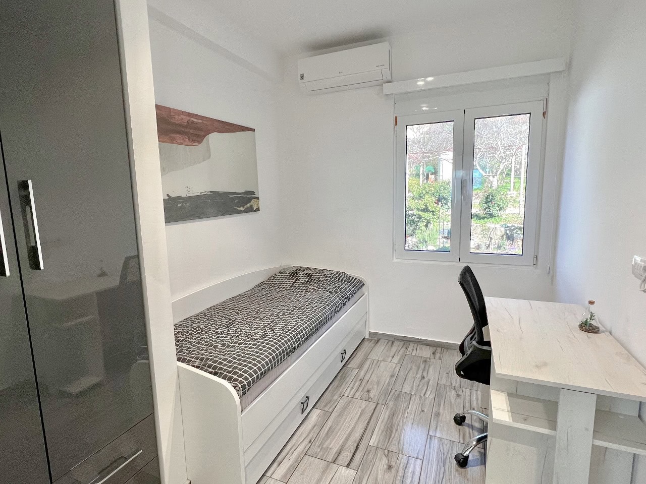 Bright bedroom with a bed, desk, and wardrobe in an apartment in Tivat, Montenegro. View of a green garden from the window.