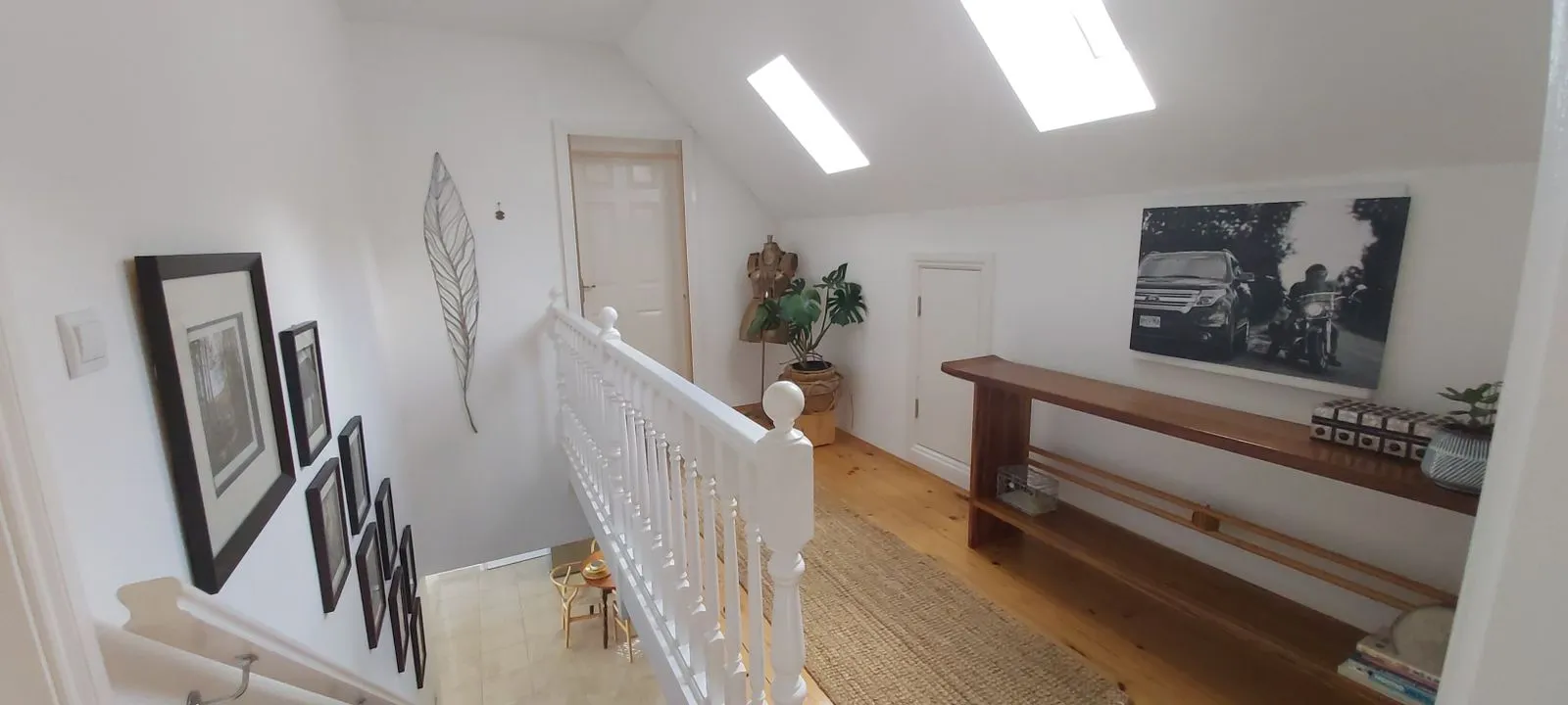 Upper landing with white railing, wood floor, long shelf, framed art, and skylights in a sloped attic ceiling.