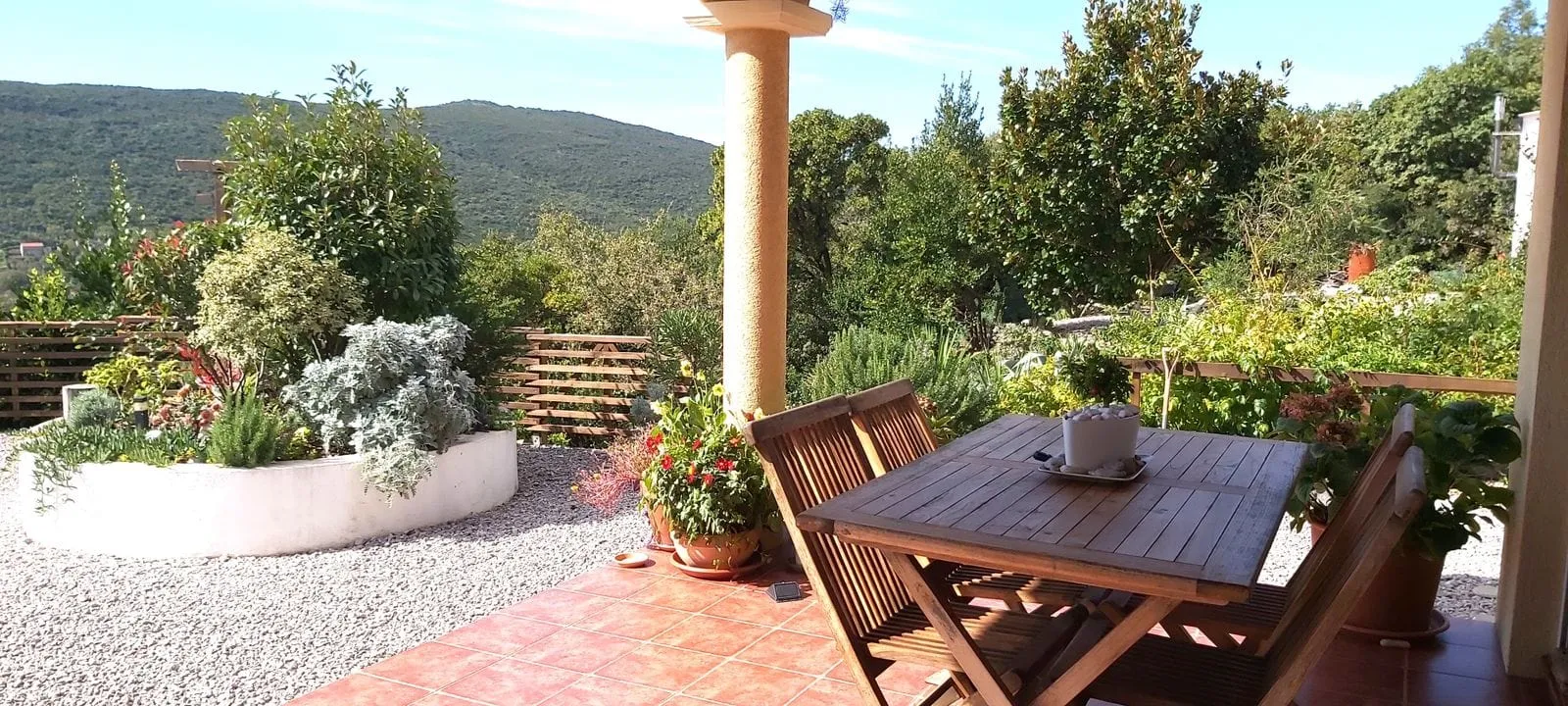 Terrace with wooden table and chairs overlooking a garden and hills.