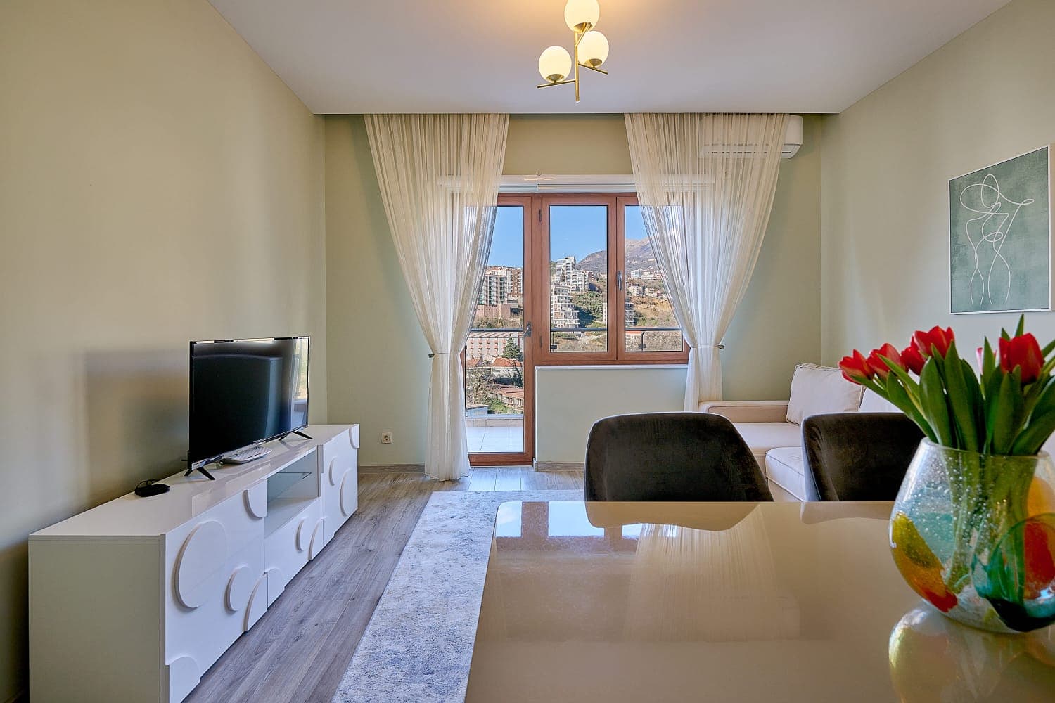 Cozy living room in an apartment in Bečići, Montenegro: light-colored furniture, TV area, and access to a balcony with mountain views.